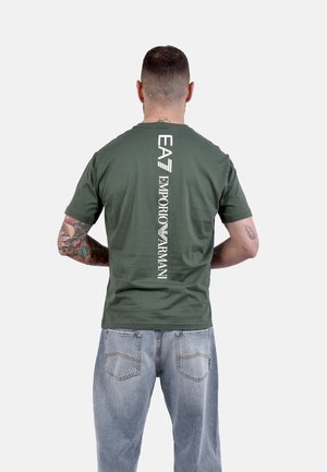 Uomo con tatuaggi che indossa una t-shirt verde EA7 Emporio Armani e jeans blu, di spalle su uno sfondo chiaro e uniforme.