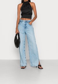 Černý halter top v kombinaci s bledě modrými širokými džínami s postranními rozparky. Modelka drží černou prošívanou kabelku a má na sobě sandály se čtveratou špičkou.