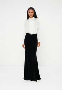 Saia maxi de veludo preto com um corte justo e um cinto com acabamento em tom dourado. Combinada com uma blusa branca de mangas compridas e colarinho.