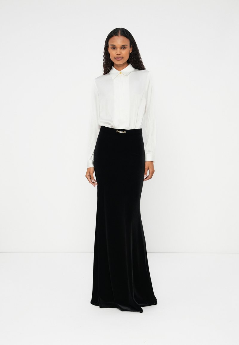 Saia maxi de veludo preto com um corte justo e um cinto com acabamento em tom dourado. Combinada com uma blusa branca de mangas compridas e colarinho.