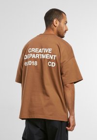Brun oversized t-shirt lavet af bomuld med hvid tekst "CREATIVE DEPARTMENT ©2018 CD" på ryggen. Korte ærmer, afslappet pasform.
