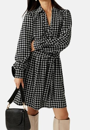 Kobieta w czarno-białej sukience w kratę z wzorem liści, trzymająca czarną teksturowaną torebkę, w połączeniu z beżowymi kozakiem za kolano.