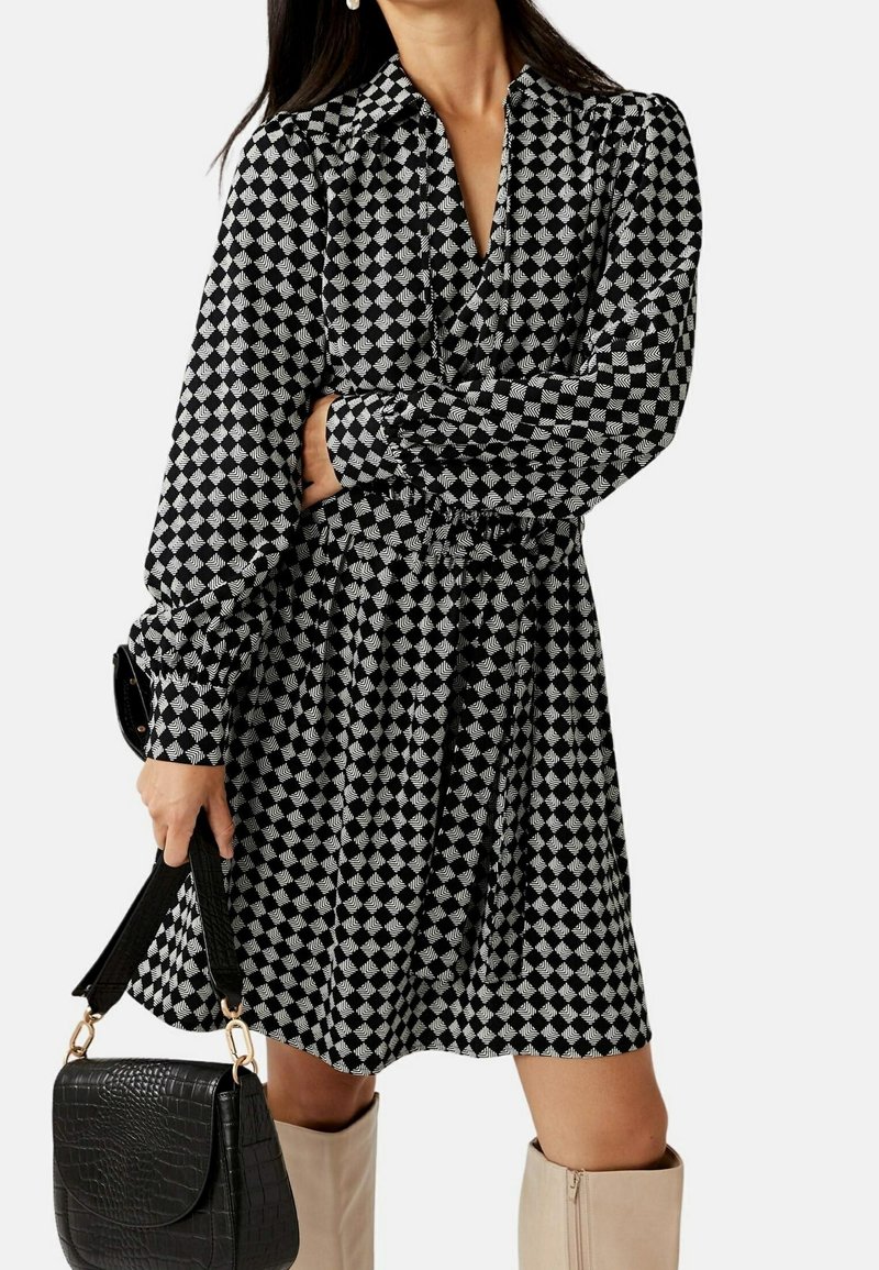 Femme portant une robe à carreaux noir et blanc avec un motif de feuilles, tenant un sac à main texturé noir, assortie à des bottes beige montant jusqu'aux genoux.
