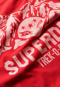 Tissu rouge avec un graphisme blanc estompé représentant un blason, des montagnes, et le mot « SUPERDRY » avec le texte « TREK-O-RIDE » en dessous.