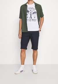 Chemise manches courtes vert olive portée sur un t-shirt blanc avec un motif de palmiers, associée à un short bleu marine à pois et des baskets blanches.