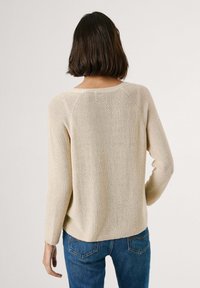 Beige neulepaita, jossa on pitkät hihat ja pyöreä pääntie. Paita on hienovaraisella tekstuurilla ja raglan-hihoilla, yhdistettynä sinisiin farkkuihin.