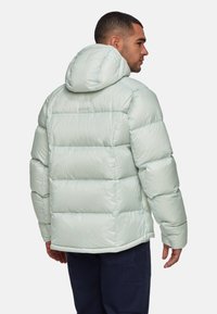 Man in een lichtgrijze gewatteerde hoodie en donkere broek, van achteren en zijkant weergegeven tegen een witte achtergrond.