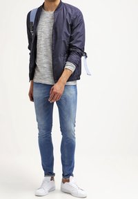 Giubbotto bomber blu navy con strisce azzurre, t-shirt grigia a righe, jeans skinny azzurri e scarpe da ginnastica bianche. Design semplice e informale.