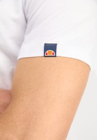 Weißes Kurzarmhemd mit einem marineblauen Etikett und einem kreisförmigen orange-roten Logo auf dem linken Ärmel, das aus weichem Stoff und glatter Textur gefertigt ist.