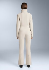 Beżowy dopasowany kombinezon z wysokim kołnierzem, długimi rękawami i rozszerzanymi spodniami z zamkami błyskawicznymi przy mankietach. Gładka, teksturowana tkanina.