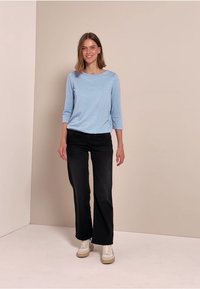 Hellblaues Oberteil mit langen Ärmeln und subtilen Glitzereffekten, kombiniert mit schwarzen Jeans und weißen Turnschuhen, vor einem neutralen Hintergrund.
