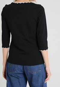 Zwarte top met driekwartmouwen en scalloped halslijn en manchetten. Zachte stof, aansluitend design, gecombineerd met blauwe spijkerbroek.
