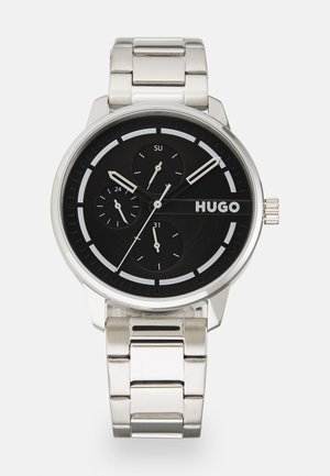 Silberner Metallzeitmesser mit schwarzem Zifferblatt, drei Subzifferblättern und fettgedruckten weißen Stundenmarkierungen. Verfügt über ein Armband aus rostfreiem Stahl und das auffällige "HUGO"-Logo.