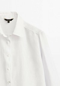 White linen shirt featuring a pointed collar, long sleeves, and five button closures in the front. Smooth texture with a lightweight design.