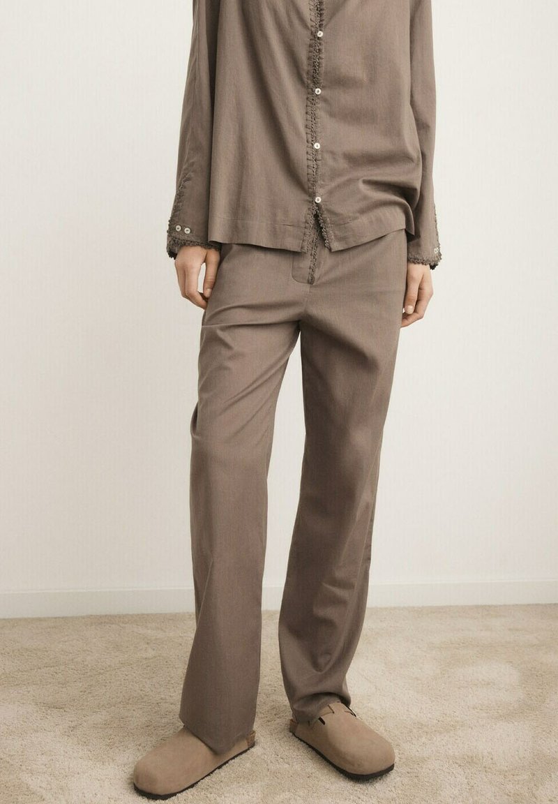 Pantalon ample marron associé à une chemise boutonnée assortie et des chaussures beige à enfiler sur un sol recouvert de moquette.