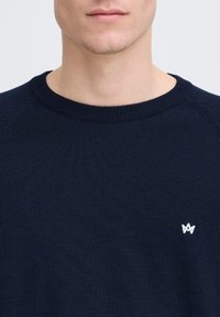 Suéter de punto azul marino con cuello redondo y puños de canalé. Presenta un pequeño logo bordado en blanco en el lado izquierdo del pecho.