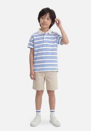 Młody chłopiec stoi, ubrany w białą koszulkę polo z niebieskimi paskami, beżowe szorty, białe skarpetki z niebieskimi paskami oraz białe trampki.