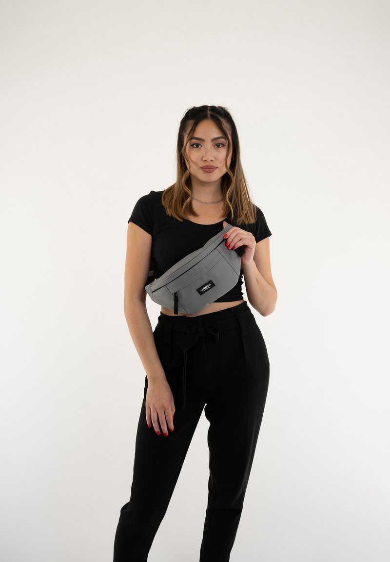 Marsupio grigio in tessuto, di forma rettangolare con un compartimento con zip, indossato a tracolla, abbinato a pantaloni neri e una maglietta nera.