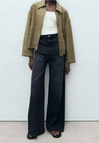Olijfgroen denim jack met knoopsluiting en voorkruiszakken, gecombineerd met hoge zwarte wijd-uitlopende jeans. Platte zwarte sandalen.