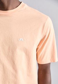 Lys ferskenfarvet kortærmet t-shirt lavet af bomuld, med et lille hvidt logo på brystet og rund halsudskæring. Glat tekstur.