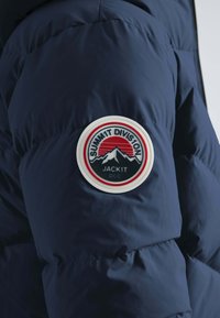 Marineblå pufferjakke med et rundt patch, der har bjerge, en sol og teksten "SUMM1T DIVISION JACK1T DCC" i rød og sort. Glat tekstur.
