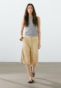 Beige suede culottes met een wijd uitlopend ontwerp, gecombineerd met een grijze mouwloze top en zwarte teenslippers, tegen een minimalistische achtergrond.