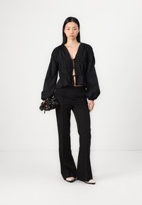 Blusa preta cortada com tecido franzido e decote em V, emparelhada com calças pretas de cintura alta. O acessório inclui uma clutch preta com padrão.