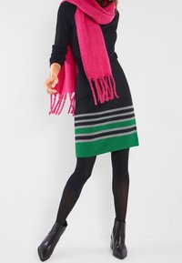 Svart klänning med grön och grårandig nederkant, kombinerad med en rosa scarf. Modellen bär svarta strumpbyxor och ankelstövlar. Mjuk textur, figurnära design.