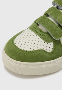 Λεπτομέρεια πράσινου σουέτ αθλητικού παπουτσιού με διάτρητο λευκό δέρμα, δύο πράσινους ιμάντες με γάντζο και βρόχο, και υφή σε εκρού καουτσούκ σόλα.