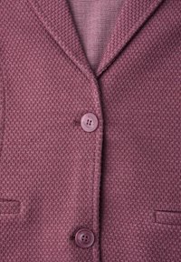 Gros plan sur un blazer rose tissé texturé avec deux boutons violets et une chemise rayée rose en dessous.