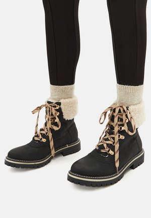 Black lace-up winter boots with thick beige shearling cuffs, beige patterned laces, worn with cream knit socks and black leggings.