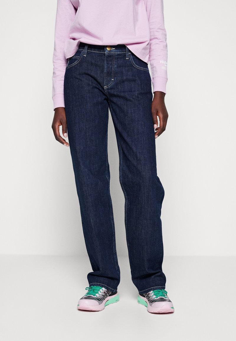 Jeans de mezclilla azul oscuro con corte recto, cinco bolsillos y costuras en contraste, combinados con zapatillas en tonos pastel y una blusa de color rosa claro.