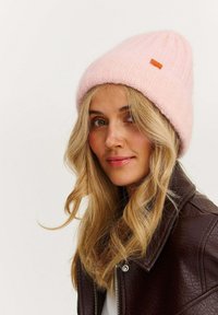 Ružová pletená čiapka s rebrovanou textúrou a malou oranžovou štítkom. Nosená s hnedou koženou bundou.