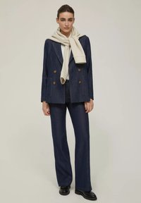 Blazer doppiopetto in denim navy con bottoni dorati abbinato a pantaloni slim-fit dello stesso colore, e una sciarpa crema chiara drappeggiata attorno al collo.