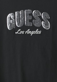 Sort stof med "GUESS" i store sølvfarvede paljetter og "Los Angeles" broderet nedenfor i hvid kursiv.