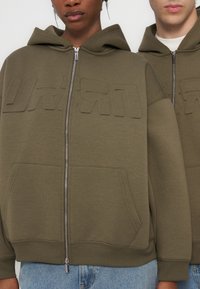 Δύο άτομα φοράνε χακί πράσινα φούτερ με κουκούλα και φερμουάρ, με ανάγλυφα γράμματα στο στήθος και μπροστινές τσέπες, που στέκονται δίπλα-δίπλα.