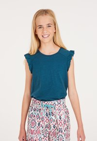 Top corto a maniche corte color teal con spalle arricciate, abbinato a pantaloni corti bianchi con coulisse e un motivo geometrico rosso e blu.