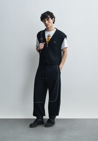 Μαύρο ριμπ αμάνικο πάνω από λευκό μπλουζάκι με γραφικά, συνδυασμένο με φαρδιά μαύρη παντελόνια με αντίθετη ραφή και μαύρα αθλητικά παπούτσια με λουράκια.