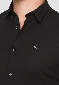 Chemise noire à boutons en tissu lisse avec col pointu. Présente un petit logo "ck" blanc brodé et des boutons assortis.
