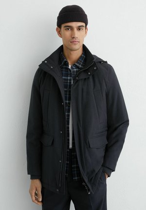 Muž v čiernej čiapke, čiernej zimnej bunde cez modrú károvanú košeľu, stojaci pred bielou stenou, tvárou vpred s neutrálnym výrazom.