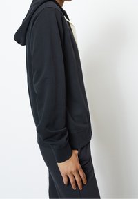 Felpa con cappuccio grigio scuro, dal tessuto morbido, con tasca a marsupio e polsini a costine, indossata con pantaloni abbinati.