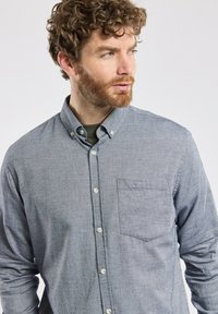 Homme aux cheveux bouclés et à la barbe, portant une chemise grise boutonnée avec poches, regardant vers sa gauche sur un fond uni.