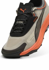 Scarpa da corsa a maglia beige con accenti neri e arancioni. Presenta una tomaia texturizzata, una suola ammortizzata e lacci riflettenti. Dettagli del logo notevoli.