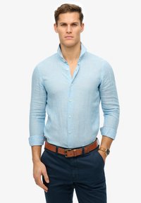 Camicia di lino azzurro chiaro con colletto a stand, chiusura con bottoni e maniche arrotolate. Indossata con pantaloni scuri e una cintura in pelle marrone.