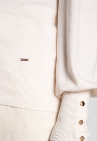 Haut en maille crème avec une texture côtelée, des accents de boutons sur la manche, et un petit détail de logo métallique près de l'ourlet.