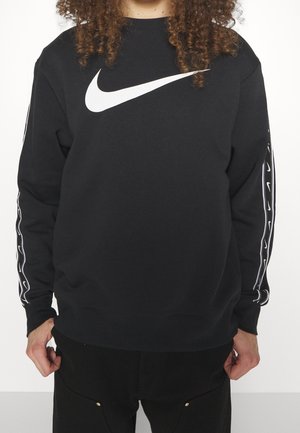 Person trägt schwarzen Nike-Sweatshirt mit großem weißem Swoosh-Logo auf der Brust und kleineren Swoosh-Mustern an den Ärmeln.
