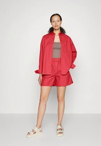 Γυναίκα στέκεται φορώντας ένα φαρδύ κόκκινο πουκάμισο με κουμπιά, κόκκινα σορτς, καφέ κοντομάνικο μπλουζάκι και λευκά σανδάλια μπροστά σε λευκό φόντο.