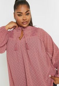 Blusa a maniche lunghe, con scollo a girocollo, di un morbido color malva con pois testurizzati, con legacci a frangia sul collo e dettagli in pizzo sulle spalle.