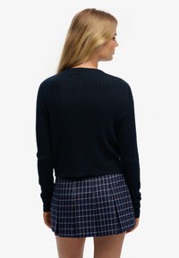 Marineblå kabelstrikket sweater med rund halsudskæring og lange ærmer, kombineret med et plisseret ternet nederdel i marineblå, rød og hvid.