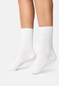 Calze di cotone bianche con bordo a coste, texture liscia e punta rinforzata, posizionate su piedi nudi su uno sfondo chiaro.